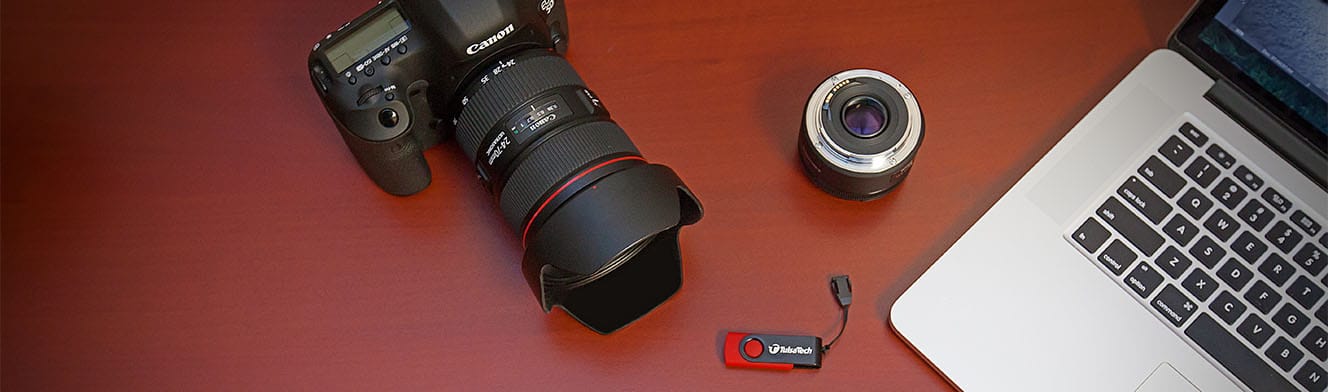 A Canon EOS 5D camera with a 24-70mm/2.8L lens, a Tulsa Tech branded USB drive, a 50mm lens, and a MacBook on a dark wood table surface.