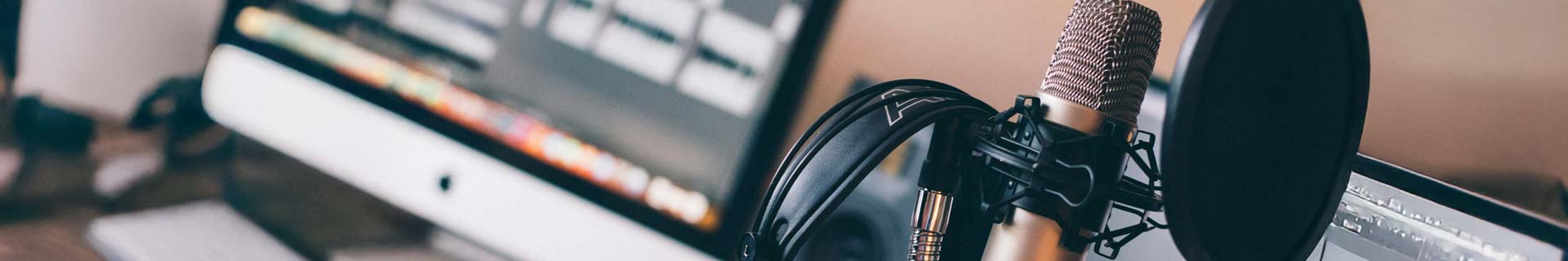 Professional microphone and headphones in the foreground with an iMac in the background with an audio editing app open.