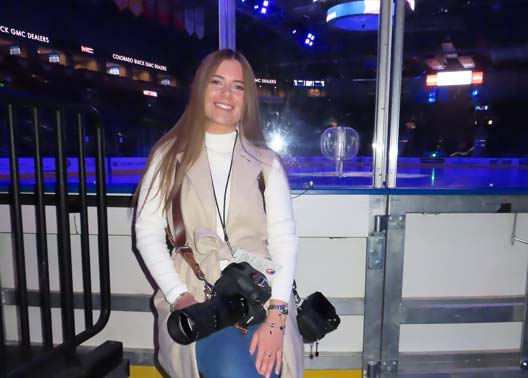 Tulsa Tech alum Kaitlyn Criss smiles while holding a camera rinkside at a hockey arena during a break from shooting.