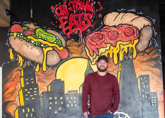 Culinary Arts graduate Max Blackstone at his Chi-Town Eats restaurant. He stands in front of mural of a large Chicago hot dog and slice of deep dish pizza over the Chicago Skyline.
