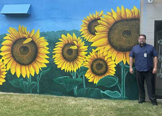 Campus Director, Dr. Sam Ramsey, stands next to the mural he created in honor of the late Becky Cunningham, a former instructor at the Children’s Learning Lab
