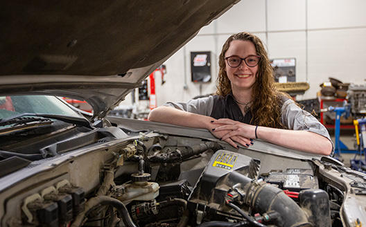 student under hood of car looking at engine smiling