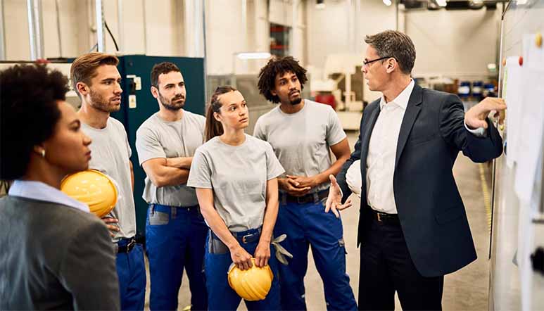 A instructor at a manuafacturing facility points at a whiteboard while talking to a group of technicians